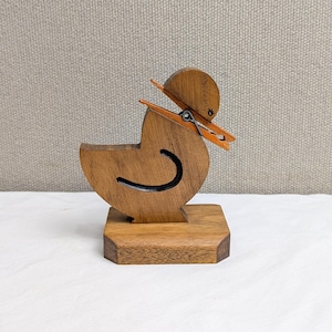 May include: Wooden duck-shaped desk organizer with a black curved line detail and an orange clothespin. The duck sits on a square wooden base. The duck's head has a small wooden hat attached with the clothespin.