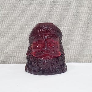 May include: A deep red translucent glass figurine shaped like the head and shoulders of Santa Claus. The piece features a textured hat, detailed facial features including a mustache and beard, and a smooth, glossy finish throughout the molded glass.