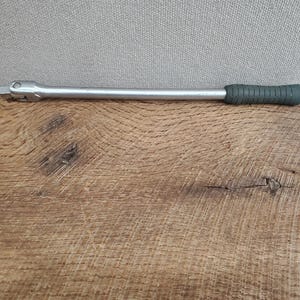 May include: A silver metal ratchet wrench with a dark green rubber grip. The wrench is lying on a wooden surface with a gray background. This tool is used for tightening or loosening bolts and nuts.