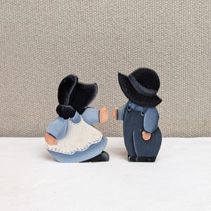 May include: Two wooden figurines of children. One is a girl in a blue dress and white apron, the other is a boy in blue overalls and a black hat. Both have black hair and are painted with simple details.