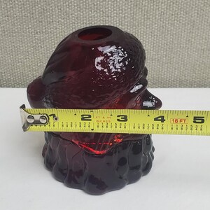 May include: A deep red glass figurine shaped like a bearded figure wearing a hat, positioned behind a yellow measuring tape that shows a width of approximately 4.5 inches. The glass has a textured surface and a hollow opening at the top.