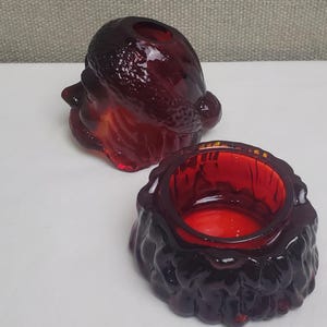 May include: A two-piece deep red glass container shaped like a Santa Claus head. The top piece features a textured hat and facial details, while the bottom piece is a circular bowl with a molded beard pattern around the exterior base.