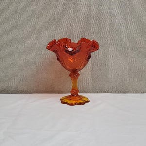 May include: An orange-colored, vintage-style glass pedestal bowl with a ruffled rim. The bowl has a detailed, textured design and sits on a slender stem and a scalloped base. The glass has a warm, amber hue.