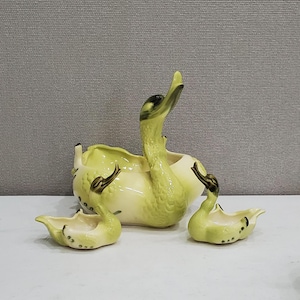 May include: A set of three green ceramic swan planters. The largest swan has its neck arched up and the two smaller swans have their necks bent down. The swans are all glazed in a light green colour with black accents.