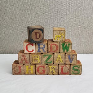 May include: A collection of vintage wooden alphabet blocks. Each block displays a different letter in various colors, including red, blue, yellow, and green. The blocks are arranged in a pyramid shape, showcasing the letters and a small illustration.