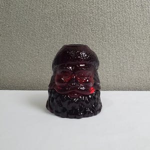 May include: A deep red glass votive candle holder shaped like the head of Santa Claus. The molded glass features a textured hat, detailed facial features including eyes, nose, and mouth, and a prominent beard covering the lower portion of the object.