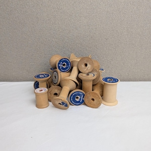 May include: A collection of light-colored wooden thread spools piled together. Several spools feature circular blue labels with white text that reads American Thread, 3 Cord Cotton, 300 Yds, and the brand name Star or Mamalicht.