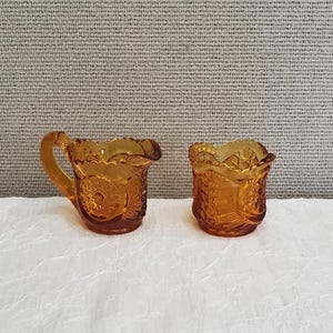 May include: A set of two amber glass creamer and sugar bowls with a decorative pattern. The bowls are made of glass and have a vintage look.