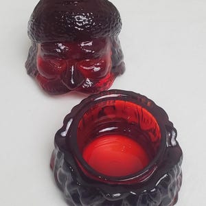 May include: A deep red glass jar with a lid shaped like a person wearing a textured hat. The base is a cylindrical container with a thick, molded rim and a smooth interior. The lid features a sculpted face with closed eyes and a textured surface.