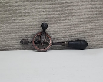 Puede incluir: Un taladro de mano vintage con un mango de madera negro y un engranaje de metal. El taladro tiene una perilla negra para girar el engranaje.