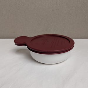 May include: A round white bowl with a deep red lid featuring a small tab handle on one side. The lid has embossed text that reads "Tupperware" along with other small, partially visible markings. The bowl has a smooth, matte finish.