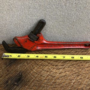 May include: Vintage red pipe wrench with black jaws and handle. The wrench is approximately 11 inches long. The text "THE RIDGE TOOL CO. ELYRIA, O." is visible on the handle. The wrench is on a wooden surface.