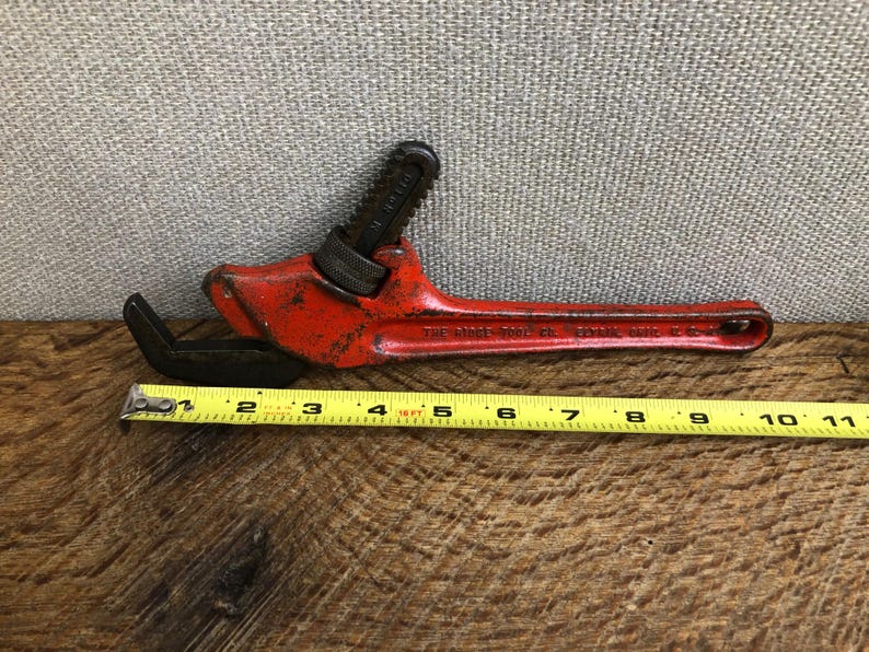 May include: Vintage red pipe wrench with black jaws and handle. The wrench is approximately 11 inches long. The text "THE RIDGE TOOL CO. ELYRIA, O." is visible on the handle. The wrench is on a wooden surface.