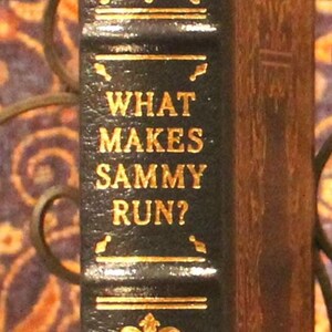 May include: A black leather-bound book with gold lettering. The title is "What Makes Sammy Run?" by Budd Schulberg. The book is a signed edition.