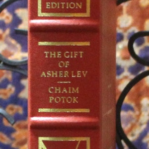 May include: A red hardcover book with gold lettering. The title is "The Gift of Asher Lev" by Chaim Potok. The book is a signed first edition and part of The Franklin Library.