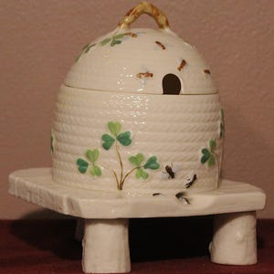 Pode incluir: Um pote de mel de cerâmica creme em forma de colmeia, decorado com trevos verdes e insetos. O pote assenta numa base retangular a condizer com quatro pernas curtas. A tampa tem uma pequena abertura.