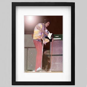 Puede incluir: Impresión enmarcada que presenta una actuación en el escenario. La imagen muestra a un músico con una chaqueta colorida y pantalones rojos, tocando una guitarra blanca. El fondo incluye amplificadores y un escenario. La impresión está en un marco negro.