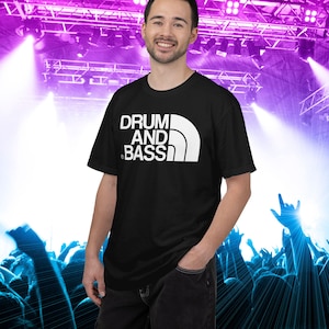 Puede incluir: Una camiseta negra de manga corta con un estampado gráfico blanco en el pecho que dice DRUM AND BASS en una fuente estilizada, acompañado de un elemento gráfico blanco curvado que recuerda a un logotipo de montaña a la derecha.