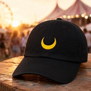 May include: A black baseball cap featuring a yellow embroidered crescent moon symbol centered on the front panel. The cap has a curved brim and a structured crown with stitched eyelets, showcasing a clean and simple design on the fabric.
