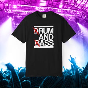 May include: A black short-sleeved t-shirt featuring a white graphic print that reads DRUM AND BASS in capital letters. The letters D and B are crossed out with a bright red X. The design is framed by two horizontal white lines above and below.