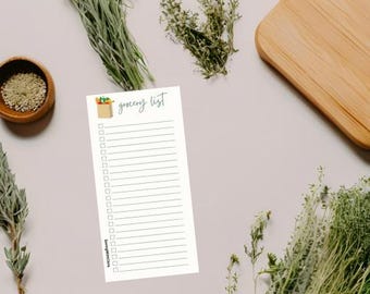 Puede incluir: Un bloc de notas blanco con la inscripción "grocery list" en verde y una pequeña ilustración de comestibles. El bloc de notas está rodeado de hierbas, un cuenco de madera y una tabla de cortar de madera.