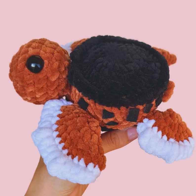Puede incluir: Un peluche de tortuga hecho a mano. La tortuga presenta una cabeza y un cuerpo marrones, un caparaz&oacute;n negro y aletas con bordes blancos. El peluche tiene un ojo negro grande y est&aacute; hecho con hilo suave y texturizado.