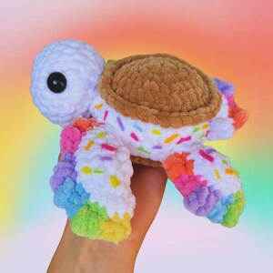 Puede incluir: Una tortuga de ganchillo hecha a mano con cabeza y cuerpo blancos, caparaz&oacute;n marr&oacute;n y patas arco&iacute;ris. La tortuga est&aacute; decorada con chispas de colores. El juguete se sostiene en una mano sobre un fondo colorido.