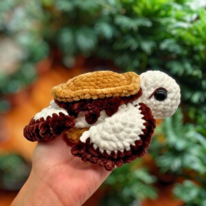 Puede incluir: Una tortuga de ganchillo hecha a mano en tonos marr&oacute;n, crema y blanco. La tortuga tiene un caparaz&oacute;n texturizado, ap&eacute;ndices blancos en forma de aleta y una cabeza color crema con un ojo negro. La tortuga se sostiene en una mano.