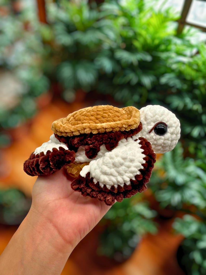 Puede incluir: Una tortuga de ganchillo hecha a mano en tonos marr&oacute;n, crema y blanco. La tortuga tiene un caparaz&oacute;n texturizado, ap&eacute;ndices blancos en forma de aleta y una cabeza color crema con un ojo negro. La tortuga se sostiene en una mano.
