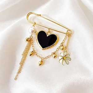 May include: A gold-toned safety pin brooch featuring a central black heart charm, a white pearl bead, a small white daisy flower charm, and several tiny gold heart charms suspended from delicate gold chains and a dangling chain fringe.