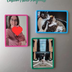 May include: Three rectangular custom photo magnets with coloured frames. One pink-framed magnet features a person holding a heart. A blue-framed magnet shows two cats. A green-framed magnet displays a couple in front of a building. Text reads "Custom Photo Magnets!"