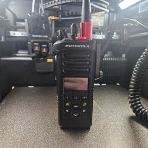 May include: A black Motorola two-way radio with a screen and buttons. The radio has a red antenna tip and is mounted inside a device. The word "MOTOROLA" is printed on the front of the radio.
