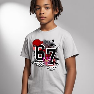 May include: A light gray t-shirt featuring the number "67" in black with a white outline, a red baseball cap, and a graphic design. The shirt also has a design of two hands and a logo.