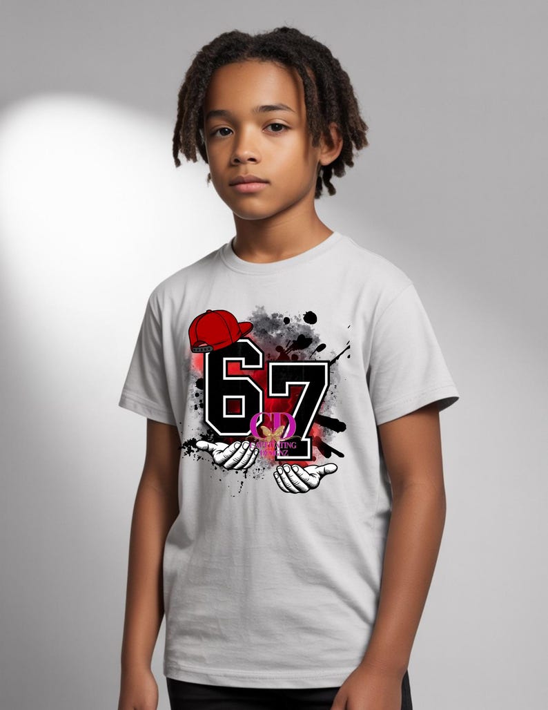 May include: A light gray t-shirt featuring the number "67" in black with a white outline, a red baseball cap, and a graphic design. The shirt also has a design of two hands and a logo.