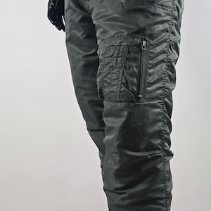 May include: Dark green cargo trousers with a drawstring waist and a zipped pocket on the leg. The trousers have a slightly shiny finish and are worn by a mannequin. Part of a casual clothing collection.