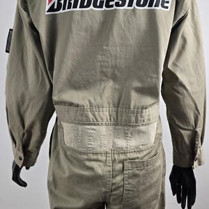 May include: Khaki-colored jumpsuit with the "BRIDGESTONE" logo in white and red on the back. The jumpsuit has long sleeves, a button-down front, and a belt detail at the waist. It appears to be made of a durable fabric.