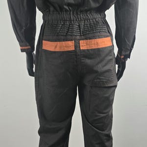 May include: Black jumpsuit with orange accents and the word "COROLLA" printed on the back. The jumpsuit features orange stripes on the sleeves and legs, and orange detailing on the back pockets.