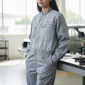 May include: A light gray mechanic's jumpsuit with the red "NISSAN" logo on the chest. The jumpsuit has long sleeves, multiple pockets, and a button-up front. The person is standing in a garage setting.