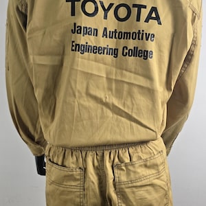 May include: Tan Toyota Japan Automotive Engineering College jumpsuit. The back of the jumpsuit features the Toyota logo and text in black. The jumpsuit has long sleeves, a button-up front, and pockets on the trousers.