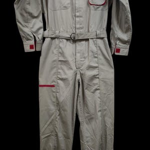 May include: Tan-colored jumpsuit with a red and white Bridgestone logo patch. The jumpsuit features a collar, long sleeves, two chest pockets, and a belt. Red accents are visible on the cuffs and pocket trim.