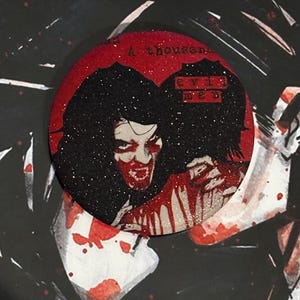 May include: A circular pin featuring a glittery red background with a graphic illustration of a person with dark hair and blood on their face and hands. The text A thousand and evil men is printed on the pin, which rests on a My Chemical Romance album cover.