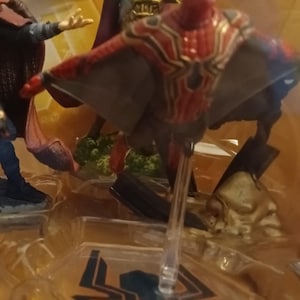 May include: A red and gold Spider-Man action figure with web wings, standing on a clear plastic base. The figure is part of a multi-figure set, with other figures visible in the background.