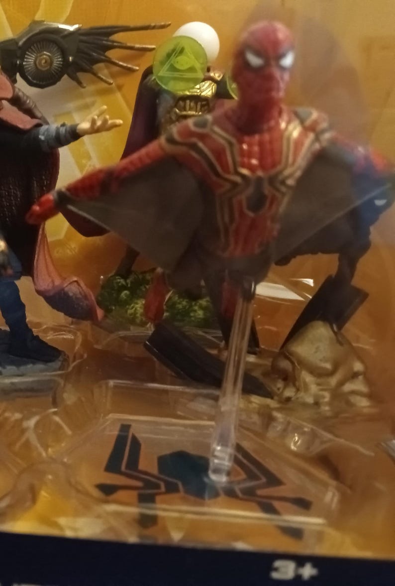 May include: A red and gold Spider-Man action figure with web wings, standing on a clear plastic base. The figure is part of a multi-figure set, with other figures visible in the background.