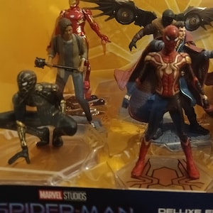 May include: A set of Marvel Studios Spider-Man: No Way Home action figures. The set includes Spider-Man in his red and black suit, Spider-Man in his black suit, Iron Man in his red and gold suit, Doctor Strange in his blue and red robe, and two other figures. The figures are all standing on a brown base with a spider web design.