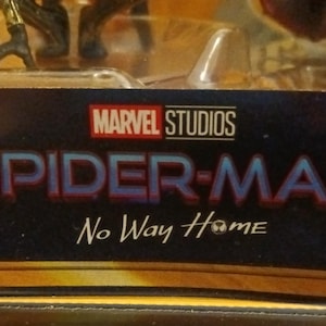 May include: A close-up of a Spider-Man No Way Home toy packaging featuring the Marvel Studios logo and the movie title. The packaging is gold and black with a blue background.