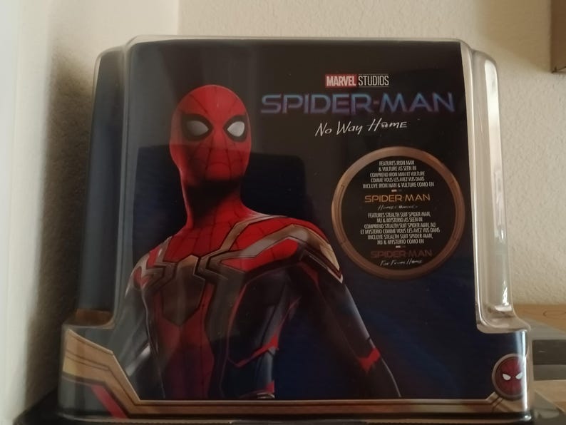 May include: A red and black Spider-Man figure in a clear plastic case with the text "MARVEL STUDIOS SPIDER-MAN No Way Home" on the front. The figure is wearing a red and black suit with a gold spider emblem on the chest.