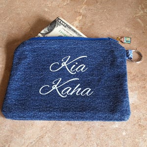 May include: A blue denim zippered pouch with the words "Kia Kaha" in white script. A dollar bill is partially visible inside the pouch. The zipper pull has a small charm and a silver key ring.