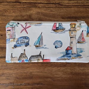 Puede incluir: Una bolsa rectangular con cremallera y temática costera. La tela blanca está estampada con ilustraciones de acuarela de veleros, un faro, cabañas de playa y una furgoneta azul. Un dije de concha cuelga de la cremallera.