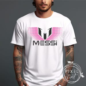 May include: White t-shirt with a pink and black graphic of a stylized "M" with the word "MESSI" underneath.