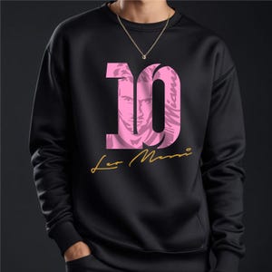 May include: Black sweatshirt with a pink number 10, featuring a silhouette of a person's face inside the number. The text "Leo Messi" is written in gold below the number.